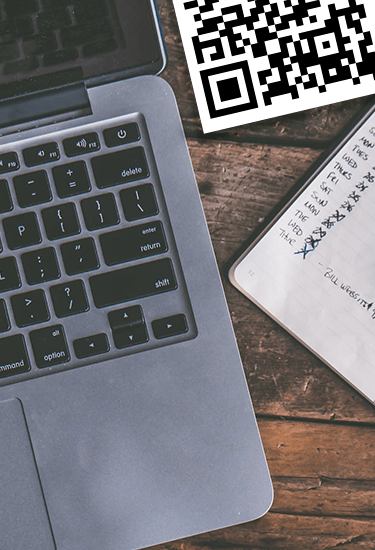 Close-up photo of an Apple Macbook Pro with a QR code on the top right and a notebook with writing on it